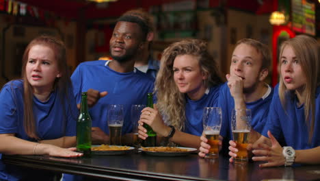 A-group-of-fans-of-men-and-women-in-blue-t-shirts-rejoice-shouting-and-drinking-beer-at-the-bar.-African-American-and-European.